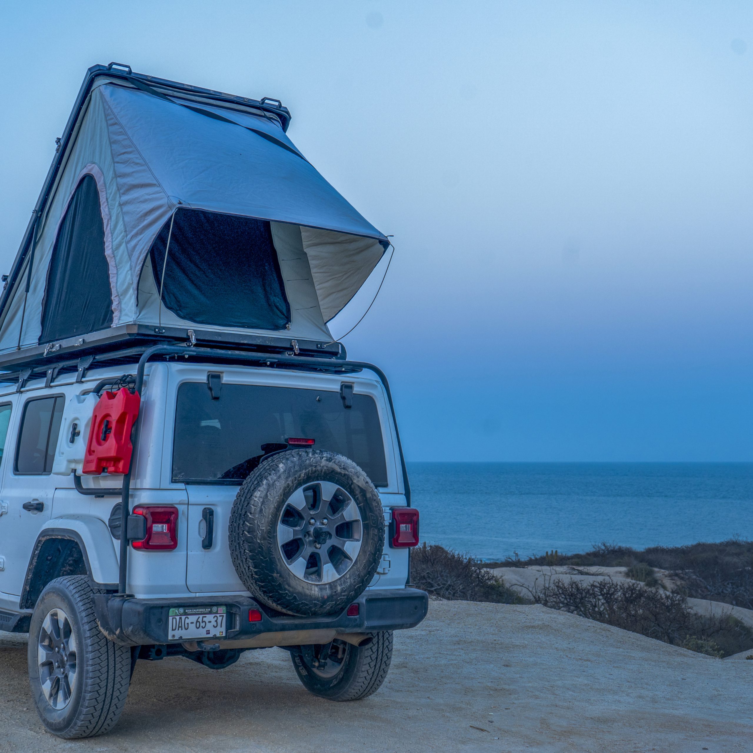 Jeep Wrangler with camper setup driving away on a sandy trail, surrounded by desert vegetation, with a scenic beach in the background.