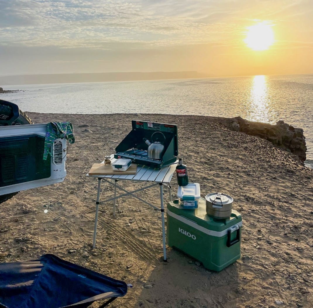 Camping Cookware setup at sunset by the sea, featuring a portable stove, cooking gear, folding table, and a cooler on rugged terrain.